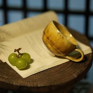 Può includere: Una tazza in ceramica fatta a mano con una glassa gialla e marrone, appoggiata su un libro aperto. La tazza ha un piccolo manico e una superficie strutturata. Accanto alla tazza ci sono tre acini d'uva verde su uno stelo. La tazza è su una superficie di legno.