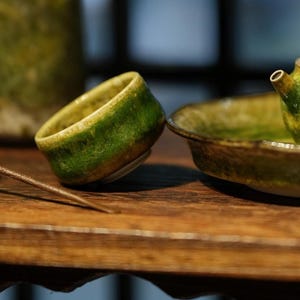 Può includere: Primo piano di un servizio da tè in ceramica con smalto verde e giallo. Il set comprende una tazzina, un piatto e una teiera. Gli oggetti sono su una superficie di legno.