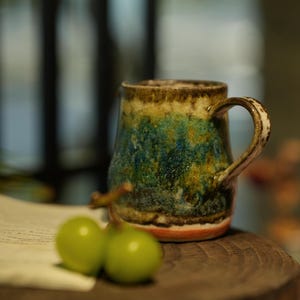 Può includere: Una tazza in ceramica fatta a mano con una smaltatura unica nei toni del blu, verde e marrone. La tazza ha un corpo arrotondato, un manico robusto e una base rosso-arancio. Due acini d'uva verde sono su una superficie di legno.