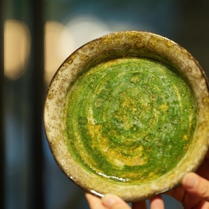 Può includere: Una ciotola in ceramica fatta a mano con un interno verde e giallo strutturato. La ciotola ha un bordo maculato dai toni terrosi e una forma poco profonda e circolare. La ciotola è tenuta da una mano.