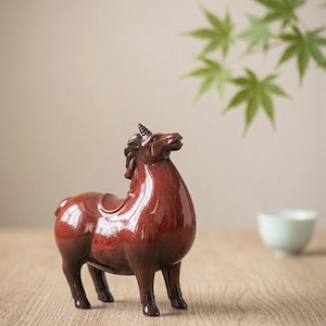 Könnte beinhalten: Eine glänzende, rotbraune Keramik-Einhornfigur. Die Fabelwesen hat eine detaillierte Mähne, ein einzelnes Horn und blickt nach oben. Die Figur steht auf einer Holzoberfläche, mit grünen Blättern und einer kleinen weißen Schale im Hintergrund.