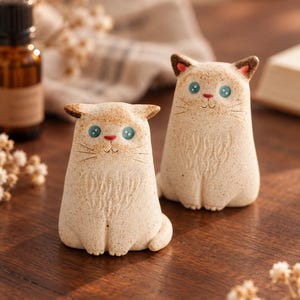 May include: Two small, beige, speckled ceramic cat figurines with pointed ears, blue eyes, and red noses. Each cat has etched details on the chest and face, including whiskers and a small mouth. The figures have a matte, textured finish.