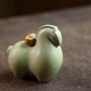 May include: A small, light green ceramic figurine of a stylized animal, possibly a sheep or a horse, with a rounded body and a small, round, tan-colored object on its back. The figurine is set against a dark, textured background.