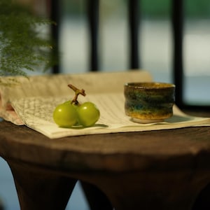 Può includere: Una piccola tazza in ceramica fatta a mano con una glassa strutturata multicolore, appoggiata su una carta vintage. Due acini d'uva verde sono vicini. I colori della tazza includono sfumature di blu, verde e oro.