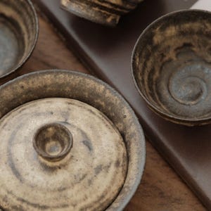 Puede incluir: Una colección de cuencos de té de cerámica hechos a mano y una tetera con tapa. Los cuencos y la tetera tienen una estética rústica y terrosa con un esmalte moteado marrón y beige. El juego de té está dispuesto sobre una superficie de madera.