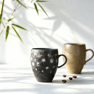 Könnte beinhalten: Zwei Keramikbecher mit Henkeln. Der vordere Becher ist dunkel mit weißen Sprenkeln, der hintere Becher ist hellbraun. Kaffeebohnen sind vor den Bechern verstreut. Bambusblätter sind im Hintergrund.