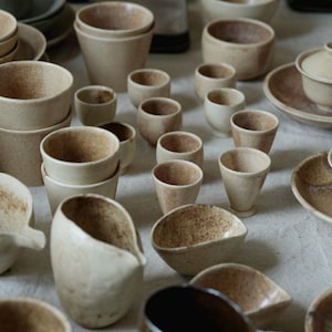 May include: An assortment of handmade ceramic tableware in various sizes and shapes. The collection includes small cups, bowls, and plates in a neutral color palette with speckled textures. The pieces are arranged on a light-colored surface.