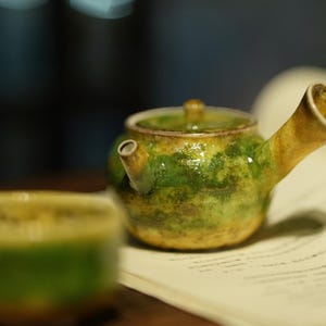 May include: A small, ceramic teapot with a green and yellow glaze, resting on a book. The teapot has a curved spout and a small handle on the lid. A matching teacup is in the foreground. The overall theme is tea and ceramics.