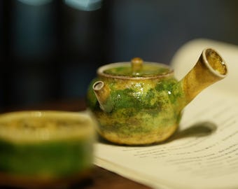 May include: A small, ceramic teapot with a green and yellow glaze, resting on a book. The teapot has a curved spout and a small handle on the lid. A matching teacup is in the foreground. The overall theme is tea and ceramics.