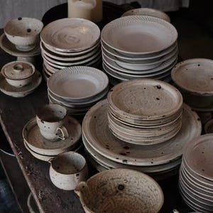 May include: A selection of handcrafted ceramic tableware, including plates, bowls, cups, and a teapot. The items are in a speckled, off-white hue with slight variations. Some plates feature a textured rim. Measurements are in centimetres.