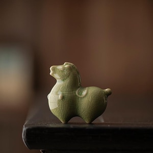 Puede incluir: Pequeña figura de caballo de cerámica verde con acabado craquelado. El caballo tiene forma redondeada, con una pequeña cola y detalle de silla. La figura se encuentra sobre un fondo oscuro y borroso.