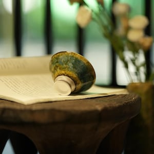 Può includere: Una piccola ciotola in ceramica con base bianca e esterno smaltato verde e oro poggia su un libro aperto. La ciotola è appoggiata su una superficie di legno scuro, con uno sfondo sfocato di verde e fiori.
