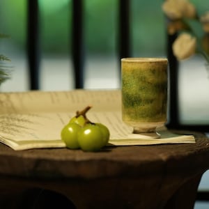 Può includere: Una natura morta con una tazza in ceramica verde e gialla, due frutti verdi e una carta arrotolata su una superficie di legno scuro. La tazza ha un design strutturato e screziato. Illuminazione soffusa e sfondo sfocato.