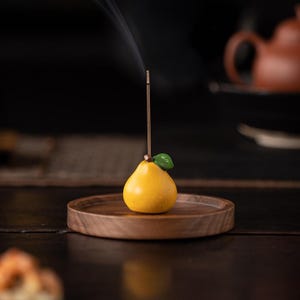 Puede incluir: Un porta incienso en forma de pera amarilla con una hoja verde, sosteniendo una varilla de incienso encendida. El soporte se encuentra sobre una bandeja redonda de madera. El humo se eleva de la varilla de incienso sobre un fondo oscuro.
