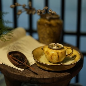 Può includere: Una piccola teiera in ceramica gialla con un piattino abbinato. La teiera ha un corpo arrotondato e un piccolo manico. Il piattino ha un bordo smerlato. Sono presenti anche un cucchiaio di legno e una piccola ciotola con foglie di tè.