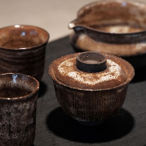 May include: A set of four brown and white ceramic teacups and a teapot. The cups and teapot have a speckled glaze and a rustic look.