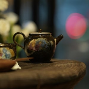 Può includere: Piccola teiera in ceramica nera con manico e beccuccio curvi. La teiera ha un coperchio ed è decorata con sfumature iridescenti blu, oro e viola. È appoggiata su una superficie di legno scuro.