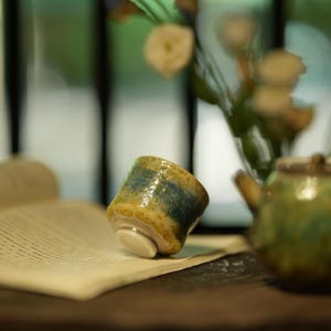 Può includere: Una piccola tazza da tè in ceramica con una glassa gialla e blu. La tazza è appoggiata su una pagina di un libro, con una teiera e dei fiori sullo sfondo. La tazza ha una base bianca.