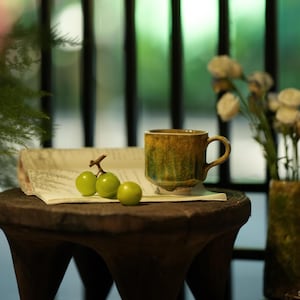 Può includere: Una tazza in ceramica con manico, nei toni del giallo, verde e marrone. La tazza è appoggiata su uno sgabello di legno, accanto a un giornale e a un grappolo di uva verde. Un vaso di fiori bianchi è visibile sullo sfondo.