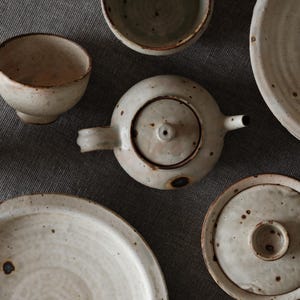 Peut inclure: Vue aérienne d'un ensemble de vaisselle en céramique faite à la main. L'ensemble comprend une théière avec couvercle, une tasse avec anse, un petit bol, une assiette et un plat couvert. Les pièces sont blanc cassé avec des mouchetures et des bords bruns.