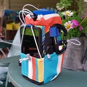 Op de afbeelding: Een kleurrijke gestreepte waterfleshouder met een telefoonvak en een karabijnhaak. De houder is gemaakt van stof en heeft een gestreept patroon in blauw, wit, rood en geel. Het telefoonvak is transparant en de karabijnhaak is zwart met de tekst "Black Diamond" erop gedrukt.