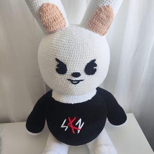 May include: A handmade crochet bunny doll. The plush toy is primarily white with long ears, peach-colored inner ears, and a black and white outfit. The black shirt features the text "4X1" in red.
