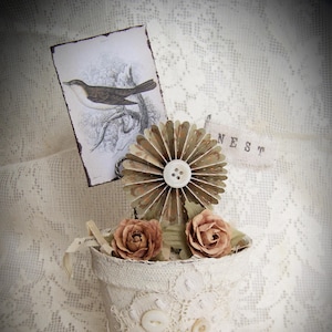 May include: A white ceramic flower pot decorated with lace and buttons. The pot is filled with artificial flowers and a paper fan. The pot has a sign that says "NEST" on it.