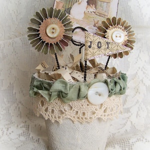 May include: A white ceramic pot with a lace trim and green fabric ruffle, decorated with paper flowers, a banner that says "JOY", and musical notes.