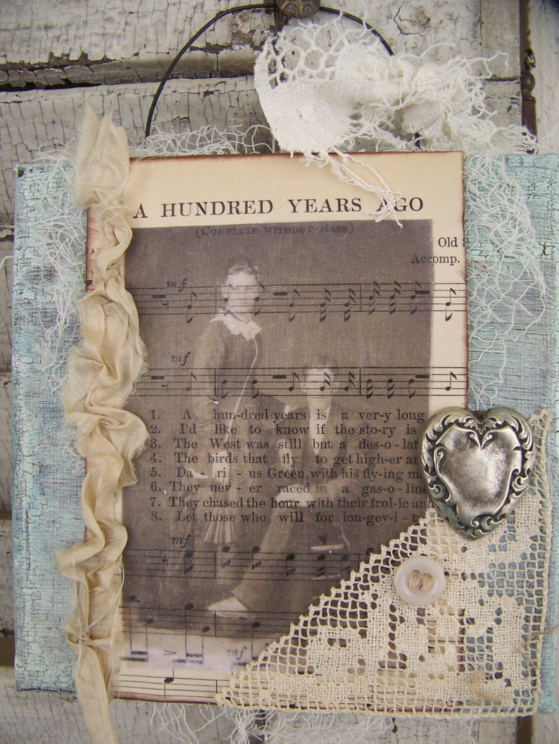 May include: A vintage-style, framed print with a musical score and lyrics for the song "A Hundred Years Ago". The print is decorated with lace, ribbon, and a silver heart charm. The text on the print reads "A Hundred Years Ago (Complete Without Bass)" and "Old Accomp."