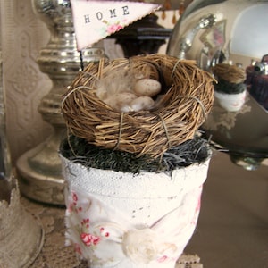 May include: A white ceramic planter with a floral design and the word "HOME" on a flag. The planter is filled with moss and a bird's nest with white eggs.