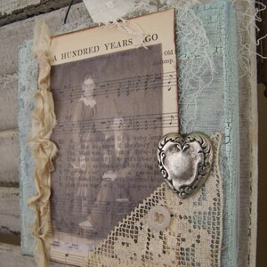 May include: A rustic wooden sign with a distressed blue paint finish. The sign features a vintage sheet music page with the text "A Hundred Years Ago" and a silver heart charm. The sign is decorated with lace and a white button.