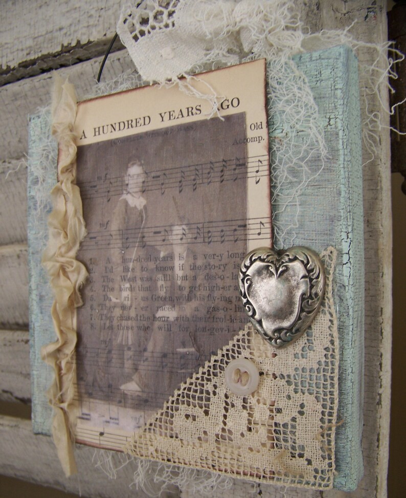 May include: A rustic wooden sign with a distressed blue paint finish. The sign features a vintage sheet music page with the text "A Hundred Years Ago" and a silver heart charm. The sign is decorated with lace and a white button.