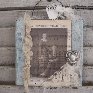 May include: A vintage-style framed artwork with a blue and white background. The artwork features a black and white photo of a young girl, a piece of lace, and a silver heart charm. The artwork has the text "A Hundred Years Ago" printed on it.