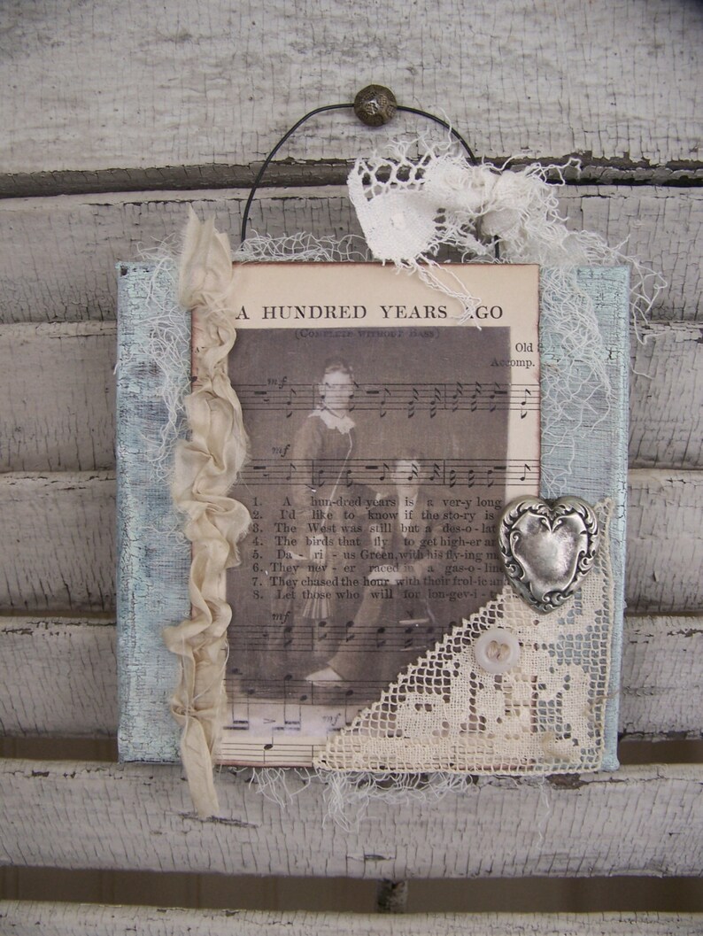 May include: A vintage-style framed artwork with a blue and white background. The artwork features a black and white photo of a young girl, a piece of lace, and a silver heart charm. The artwork has the text "A Hundred Years Ago" printed on it.