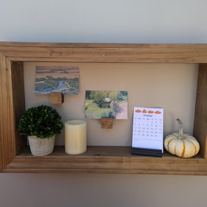 Op de afbeelding: Een houten schaduwdoos met een kleine plant in een pot, een witte kaars, twee ingelijste foto's en een kalender met de maand oktober. De kalender heeft kleine oranje pompoenen bovenaan.
