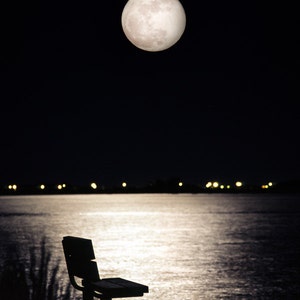 May include: A silhouette of a wooden bench sits on a shoreline with a full moon in the night sky. The water is reflecting the moonlight.