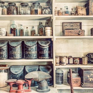 May include: A vintage general store interior with shelves stocked with various jars, bottles, and containers. The shelves are made of wood and have a white painted finish. There are several antique scales and a wooden box labeled "Cavendish Tobacco Co."