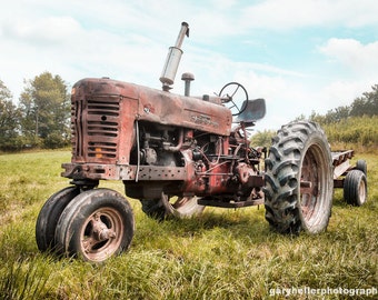 May include: A red vintage tractor with a rusted body and large tires sits in a field of green grass. The tractor has a large exhaust pipe and a small, rusted metal sign that reads "Farmall" on the side.