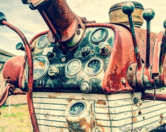 May include: Close-up of the dashboard of a vintage red tractor. The dashboard has several gauges, levers, and a white metal panel with a grid pattern. The tractor is showing signs of wear and rust.