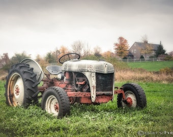 May include: A vintage, white and red tractor sits in a field of green grass. The tractor has a large, black tire on the left side and a smaller, black tire on the right side. The tractor is in a field with a house in the background.