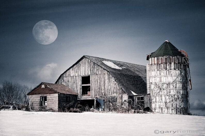 May include: A rural landscape featuring a weathered wooden barn, a silo, and a small brick building under a moonlit sky. The scene is covered in snow, creating a serene, monochromatic aesthetic. The silo has a green roof.