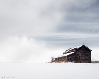May include: A weathered wooden barn stands alone in a snowy field, with a cloudy sky above. The barn has a red roof and several windows. The scene is minimalist and evokes a sense of solitude.