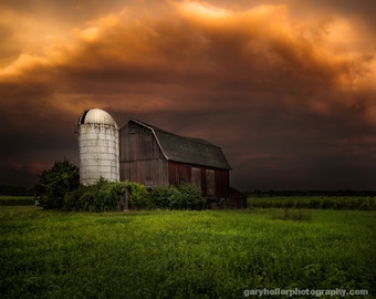 May include: A white silo and a red barn stand in a field of green grass under a dramatic, stormy sky. The sky is a mix of orange and dark gray clouds.