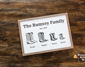 May include: A white framed sign with black text that reads "The Rumsey Family Est. 2017".  Below the text are five pairs of black and white cowboy boots with the names "Trent", "Kylie", "Vance", and "Avery" printed below each pair.