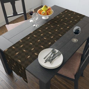 May include: A brown and tan checkered table runner with a repeating pattern of a person riding a horse. The runner is placed on a dark gray wooden table, with a white bowl of apples, wine glasses, and plates.