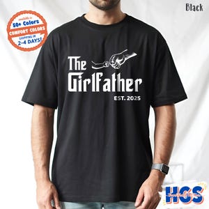 May include: Black t-shirt with the text "The Girlfather EST. 2025" in white. The design includes a graphic of two hands fist-bumping. The shirt is available in 50+ colors and made with comfort colors.