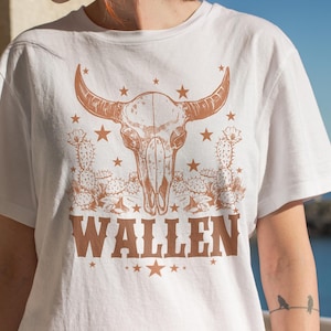 Pu&ograve; includere: T-shirt bianca con un design a teschio di bue marrone e la scritta "WALLEN" in un font in stile western. Il design include stelle e illustrazioni di cactus. Una t-shirt casual e confortevole.