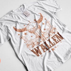 Pu&ograve; includere: T-shirt bianca con grafica di teschio di toro marrone, stelle e cactus. La parola "WALLEN" &egrave; stampata in marrone. Un'etichetta &egrave; attaccata alla maglietta.