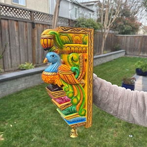 May include: A vibrantly painted, carved wooden wall bracket featuring a peacock. The bracket is primarily yellow with accents of blue, orange, green, and pink. A small gold bell hangs from the bottom. The bracket is held by a person.