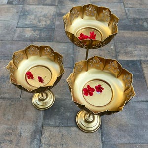 May include: Three gold-colored decorative bowls with floral cut-out edges and pedestal bases. Each bowl contains a small amount of water and a few red flower petals. The bowls are arranged on a stone surface.